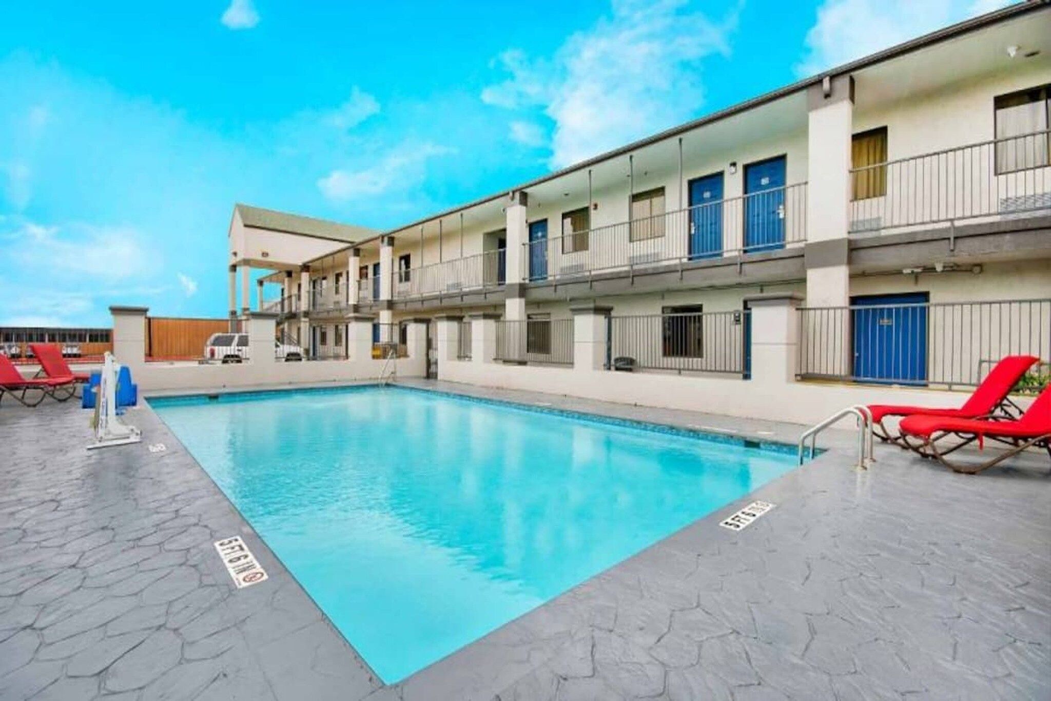 A swimming pool with a tiled deck, red lounge chairs, and a multi-story building with balconies.
