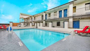 A swimming pool with a tiled deck, red lounge chairs, and a multi-story building with balconies.