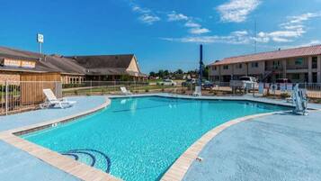 A swimming pool with a tiled deck, lounge chairs, and a building with a sign in the background.