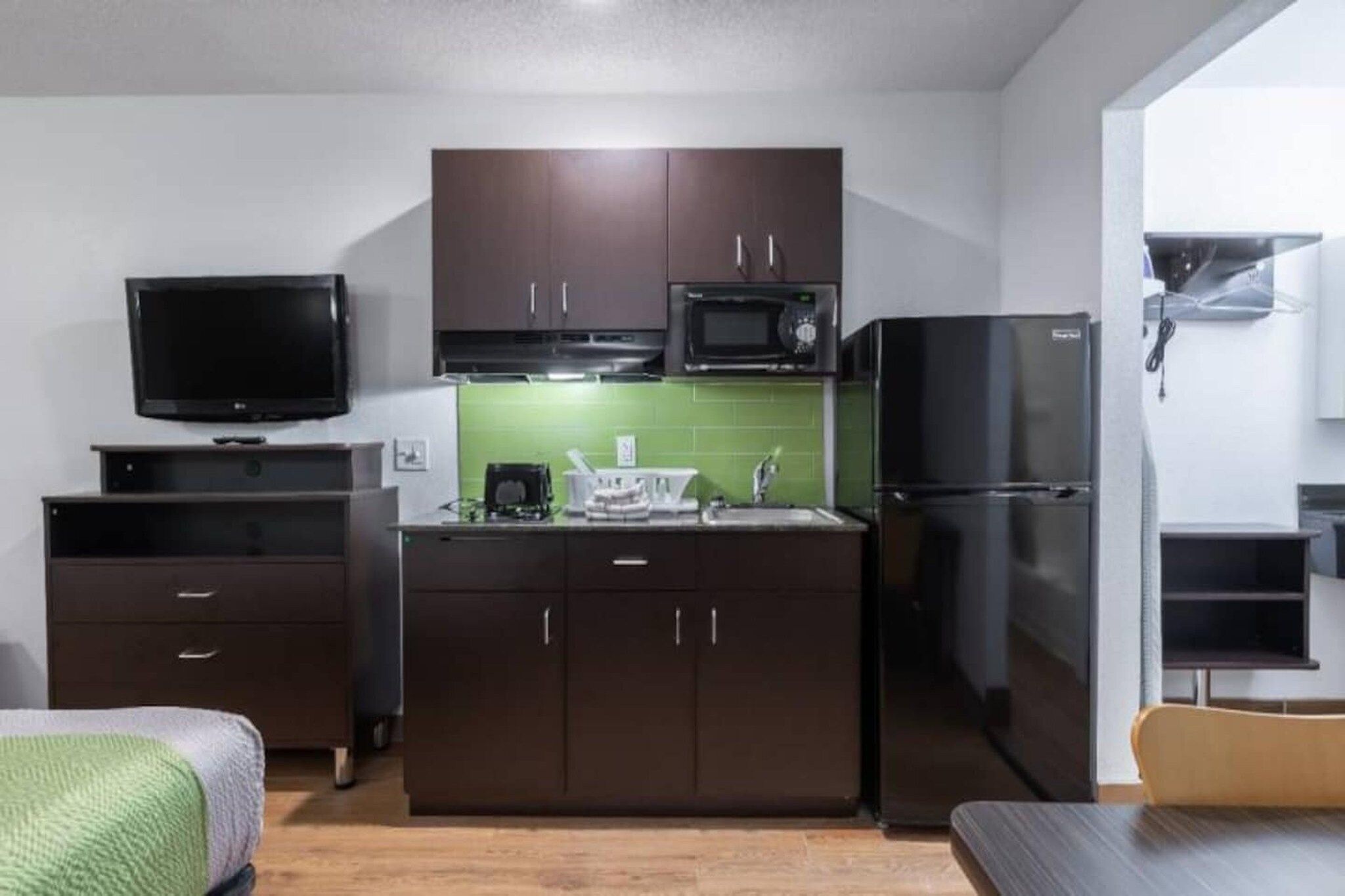 A compact kitchen with dark cabinets, a microwave, a refrigerator, a flat-screen TV, and a small dining area.