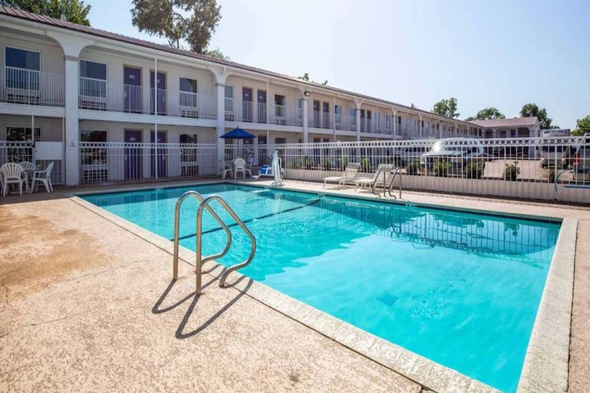 A swimming pool with a metal ladder, surrounded by a white fence, and a building with balconies in the background.