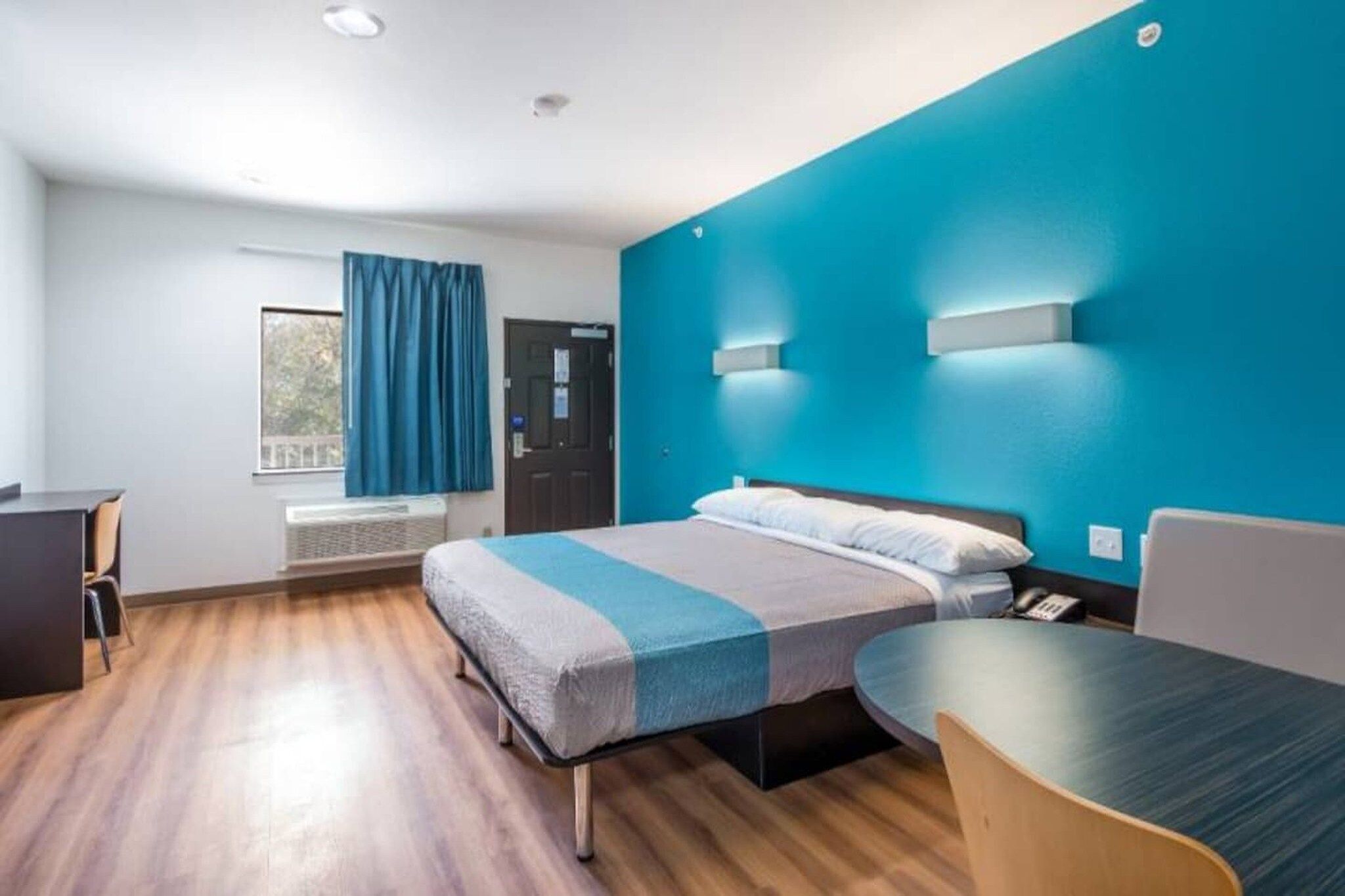 A hotel room with a bed, a desk, a chair, a window with curtains, and a wall-mounted light fixture.