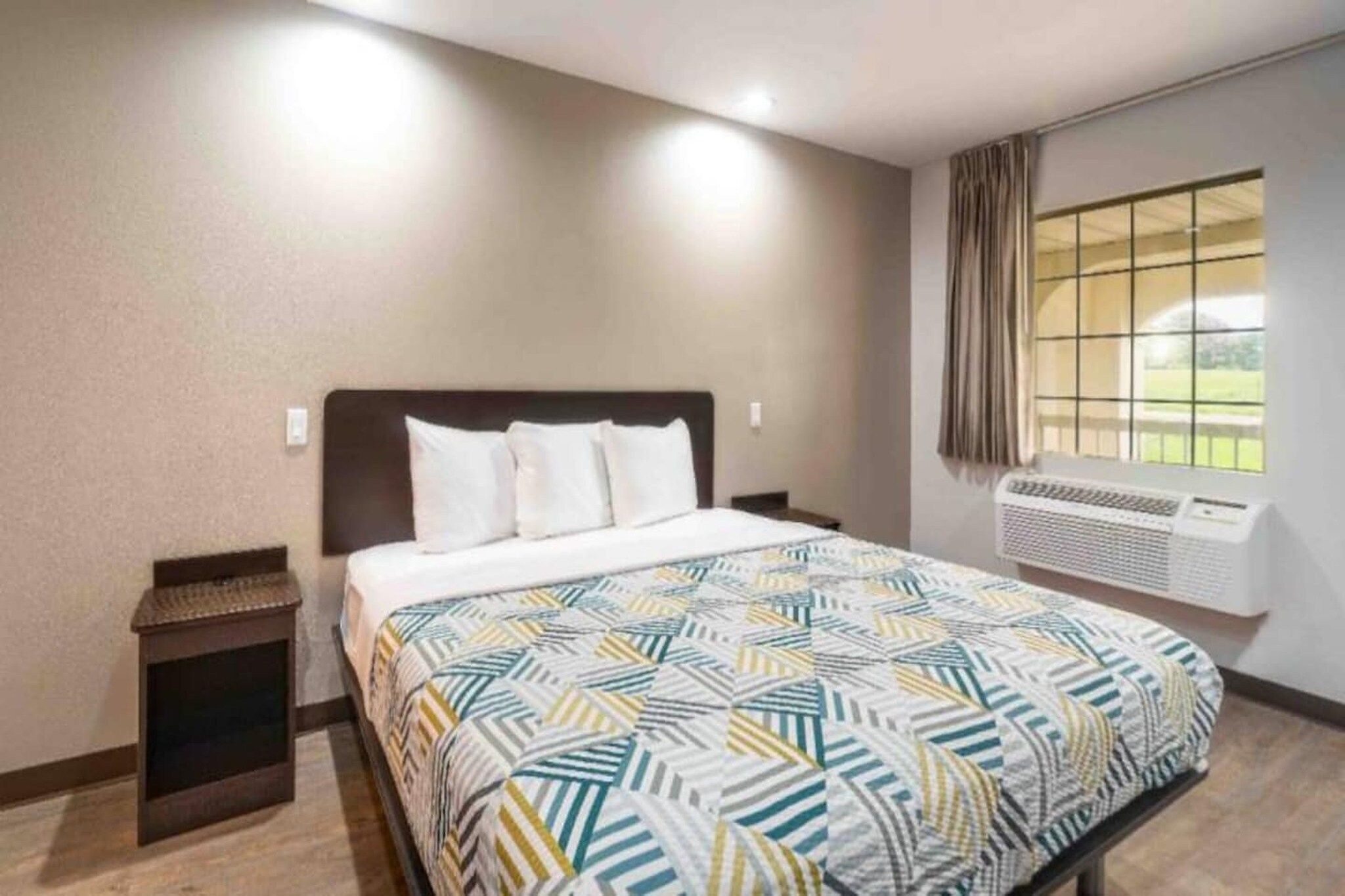 Una habitación de hotel con una cama, una mesita de noche, un ventanal con cortinas y un equipo de aire acondicionado.