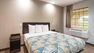 Una habitación de hotel con una cama, una mesita de noche, un ventanal con cortinas y un equipo de aire acondicionado.