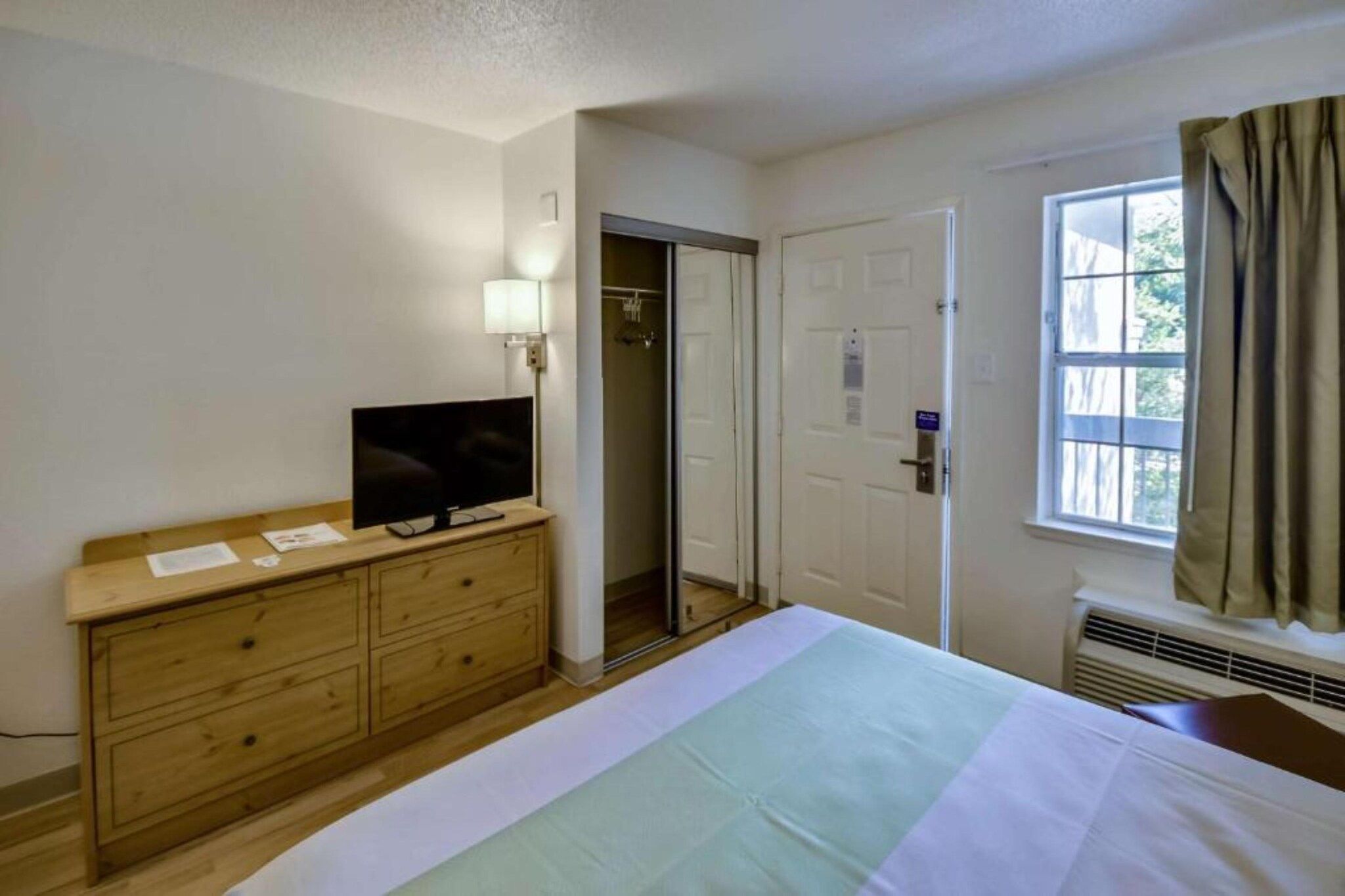 Une chambre d’hôtel avec un lit, un téléviseur posé sur une commode en bois, une fenêtre avec des rideaux et une porte donnant sur une salle de bain.