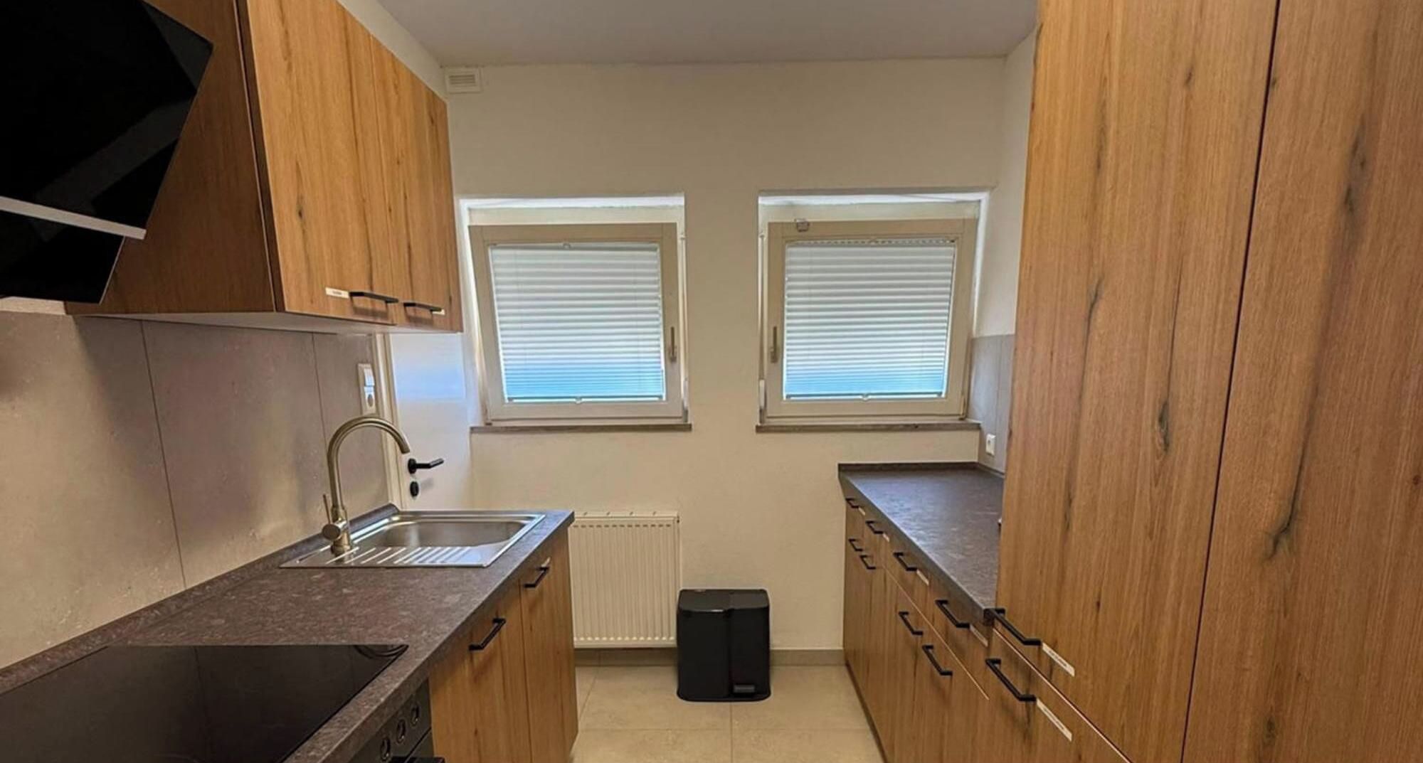 A compact kitchen with a sink, a stove, and wooden cabinets. There are two windows with blinds.