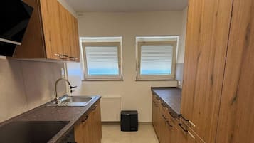 A compact kitchen with a sink, a stove, and wooden cabinets. There are two windows with blinds.