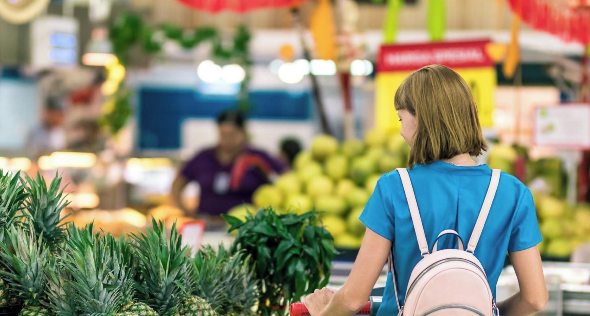 Eine Person mit Rucksack in einem Gang eines Supermarktes, mit Ananassen und anderem Obst und Gemüse.