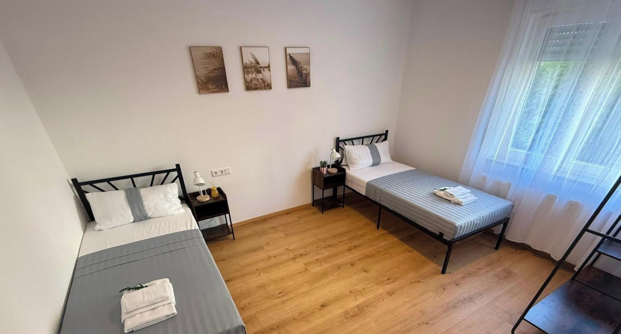 A room with two beds, a window with sheer curtains, and three wall-mounted pictures.