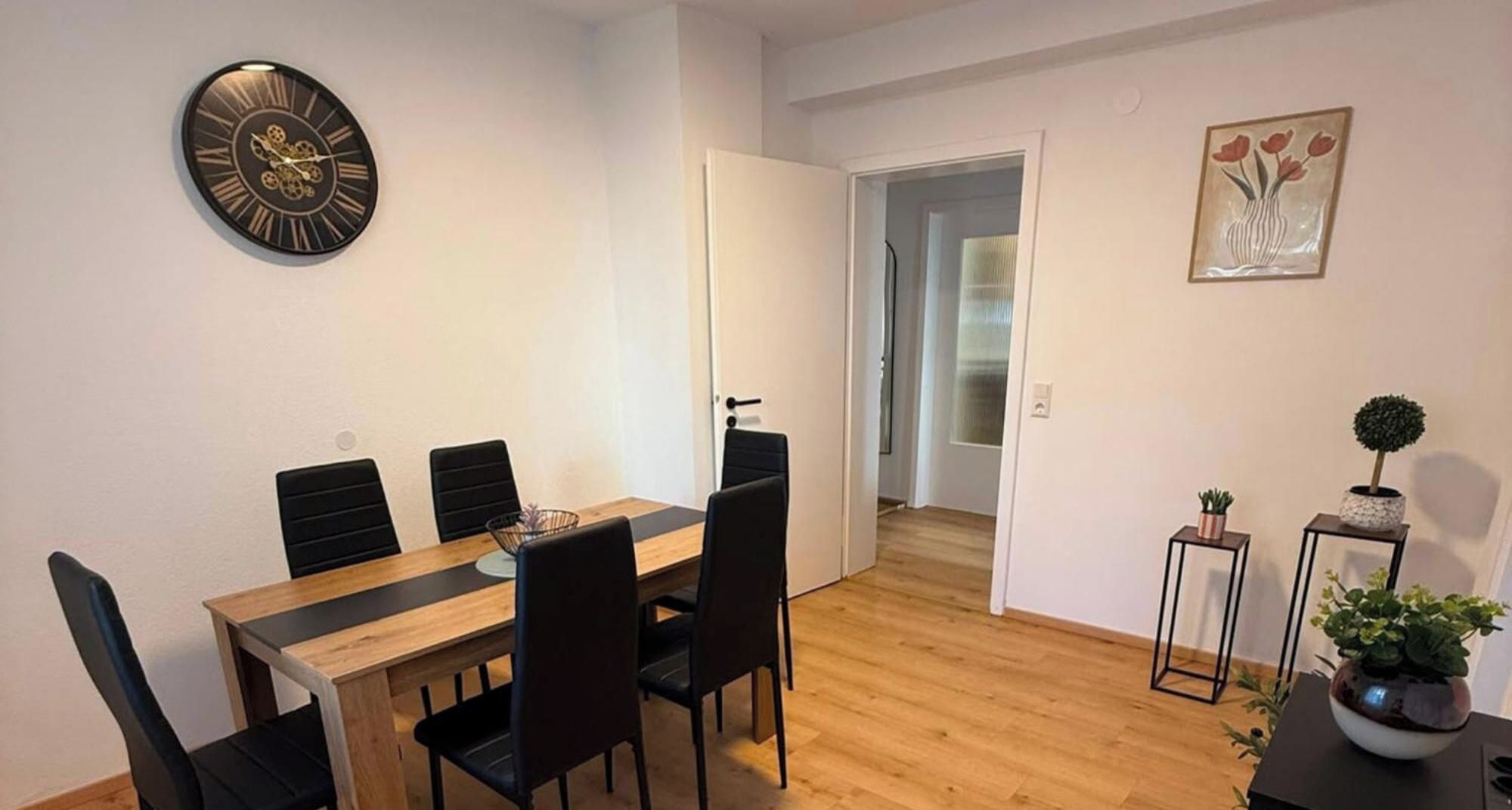 A dining area with a wooden table and black chairs, a wall clock, and a door leading to another room.