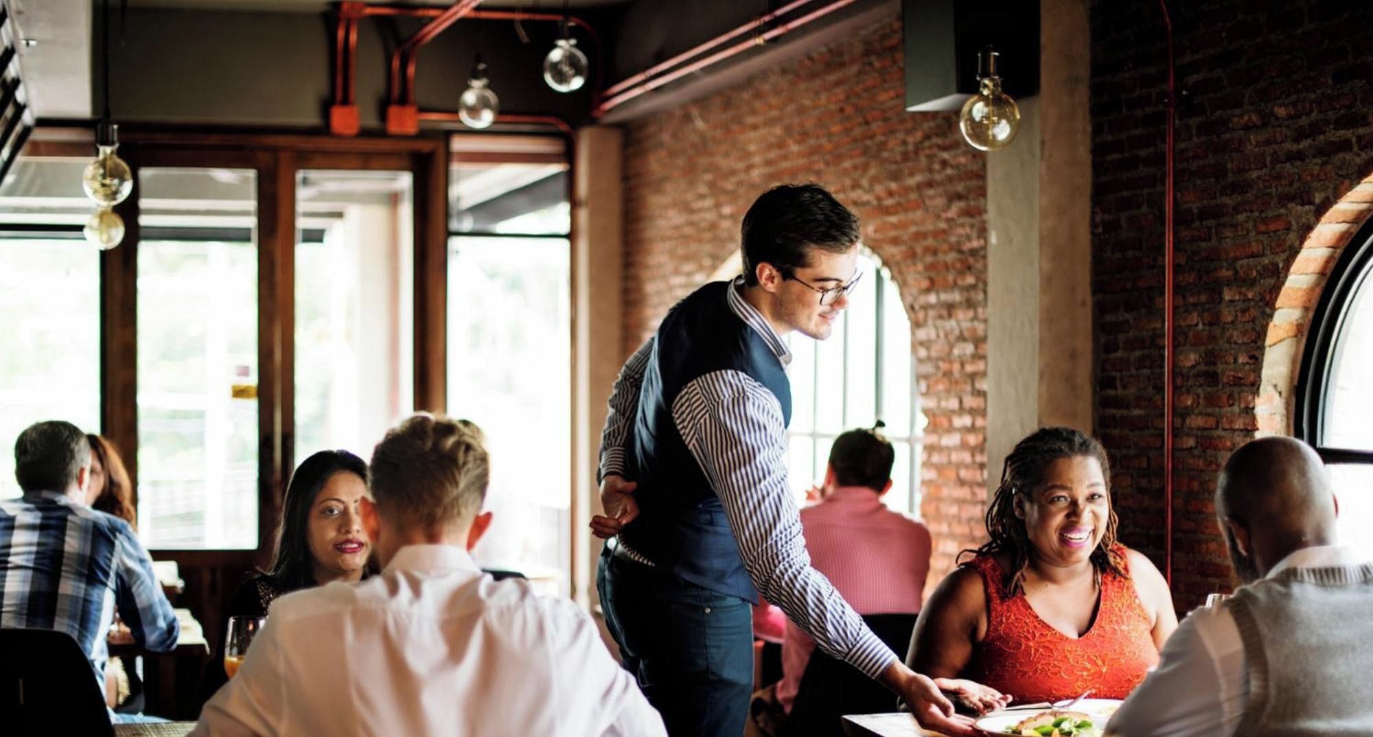 A restaurant interior with brick walls, hanging pendant lights, and patrons seated at tables.