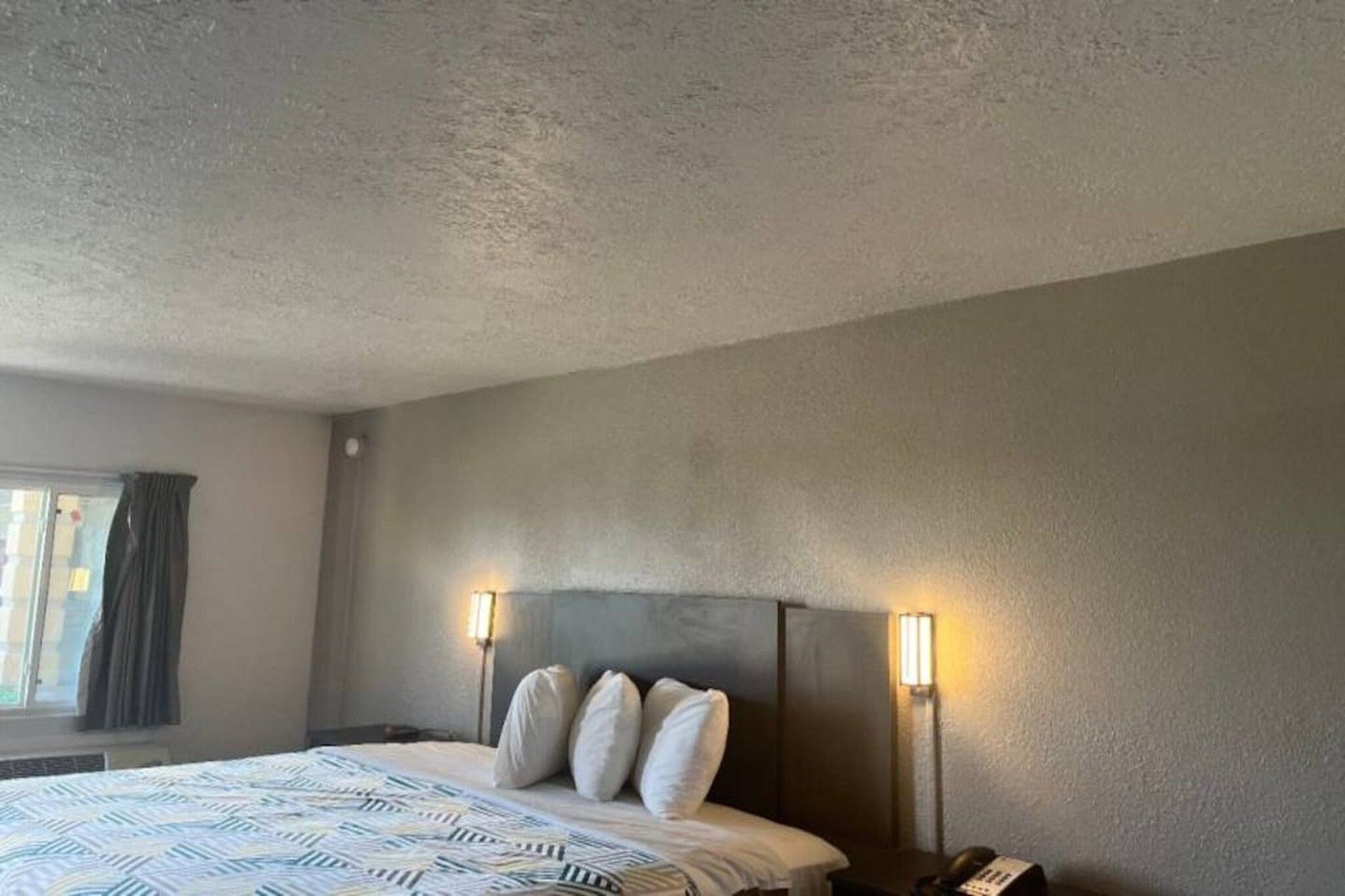 Hotellihuone, jossa on sänky, sängynpääty, yöpöytävalaisimet ja ikkuna, jossa on verhot.