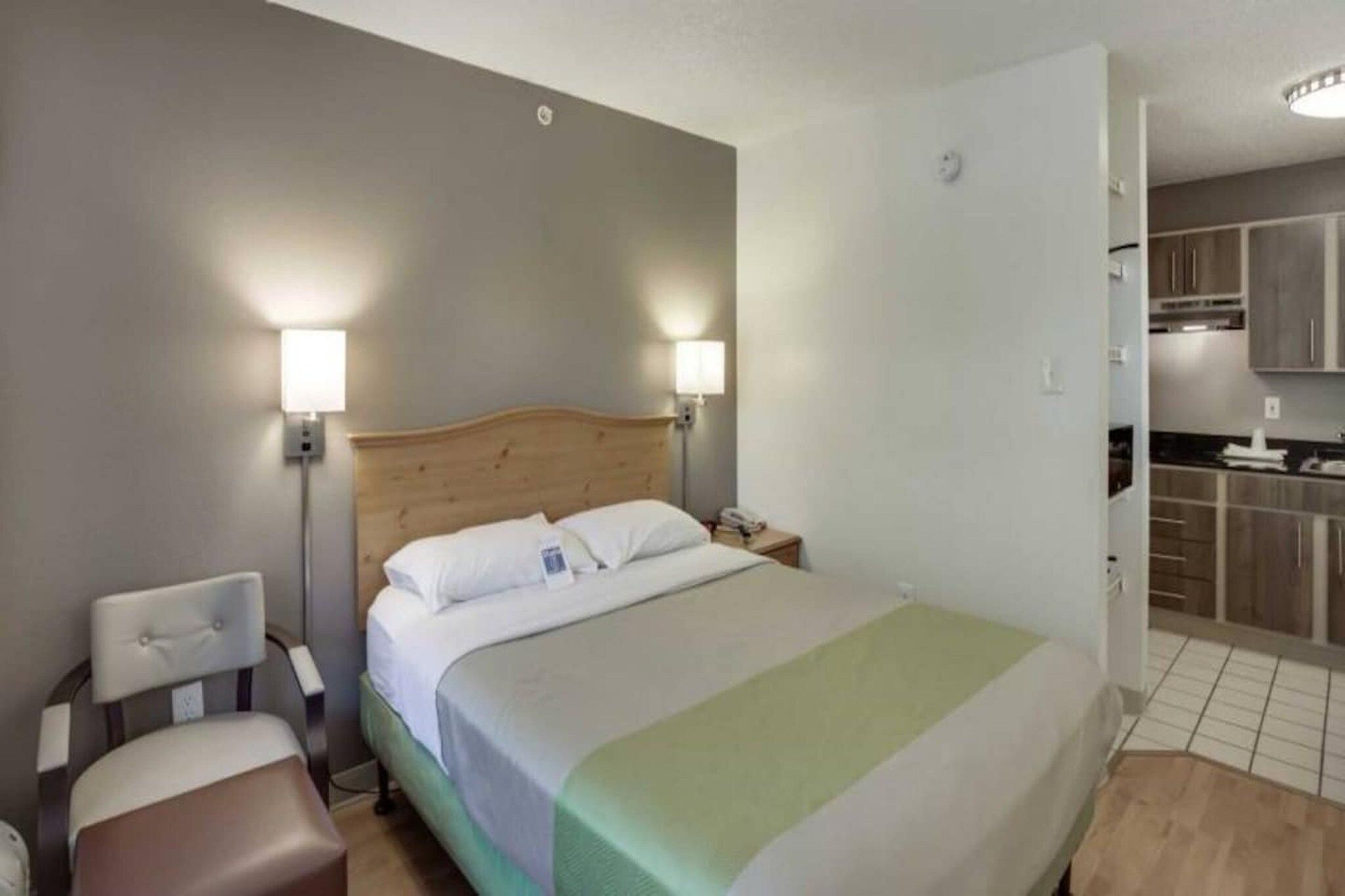 A hotel room with a bed, bedside table, lamp, chair, and a view of the kitchen.