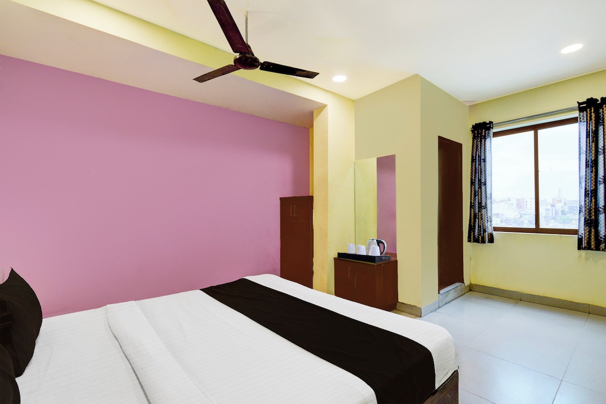 A bedroom with a bed, a ceiling fan, a window with curtains, and a small table with a kettle.