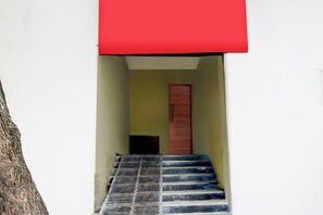 Una escalera conduce a una puerta con marco rojo, un árbol a la izquierda y una pared blanca.