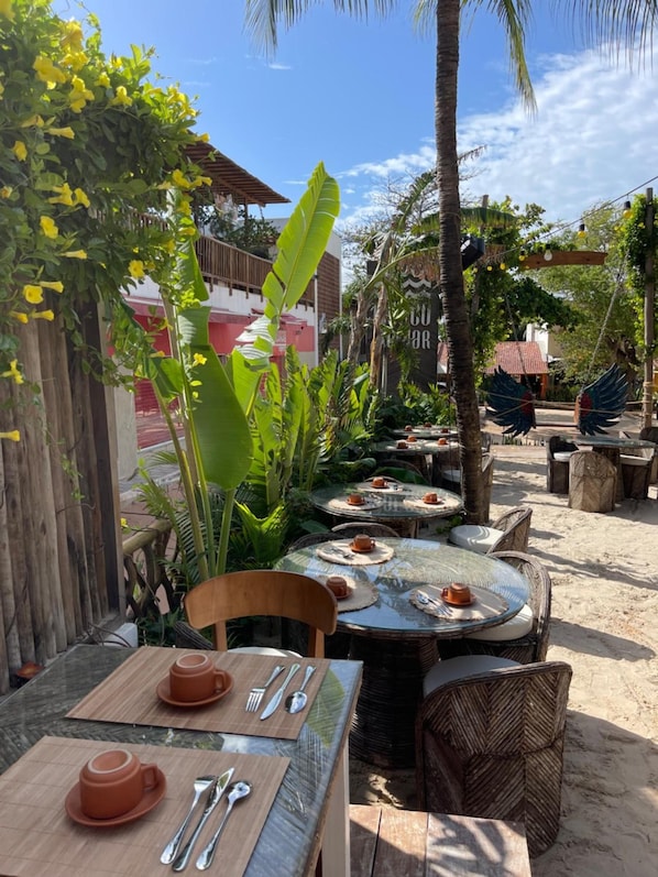 Een terras met ronde tafels en stoelen, omringd door tropische planten en palmbomen.