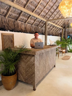 Een receptiebalie met een laptop, een grote potplant en een rieten plafond.