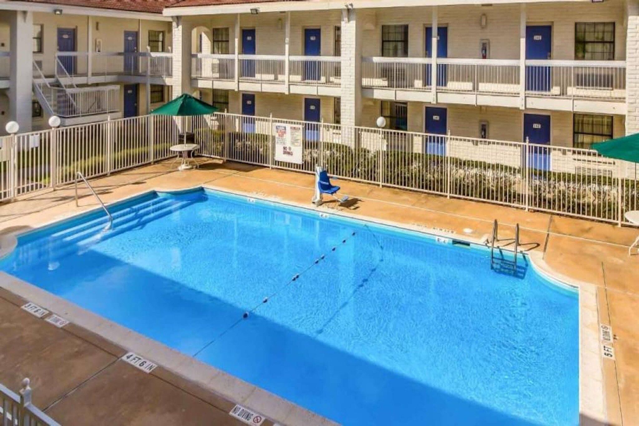 A swimming pool with a diving board, surrounded by a fenced area with a building in the background.