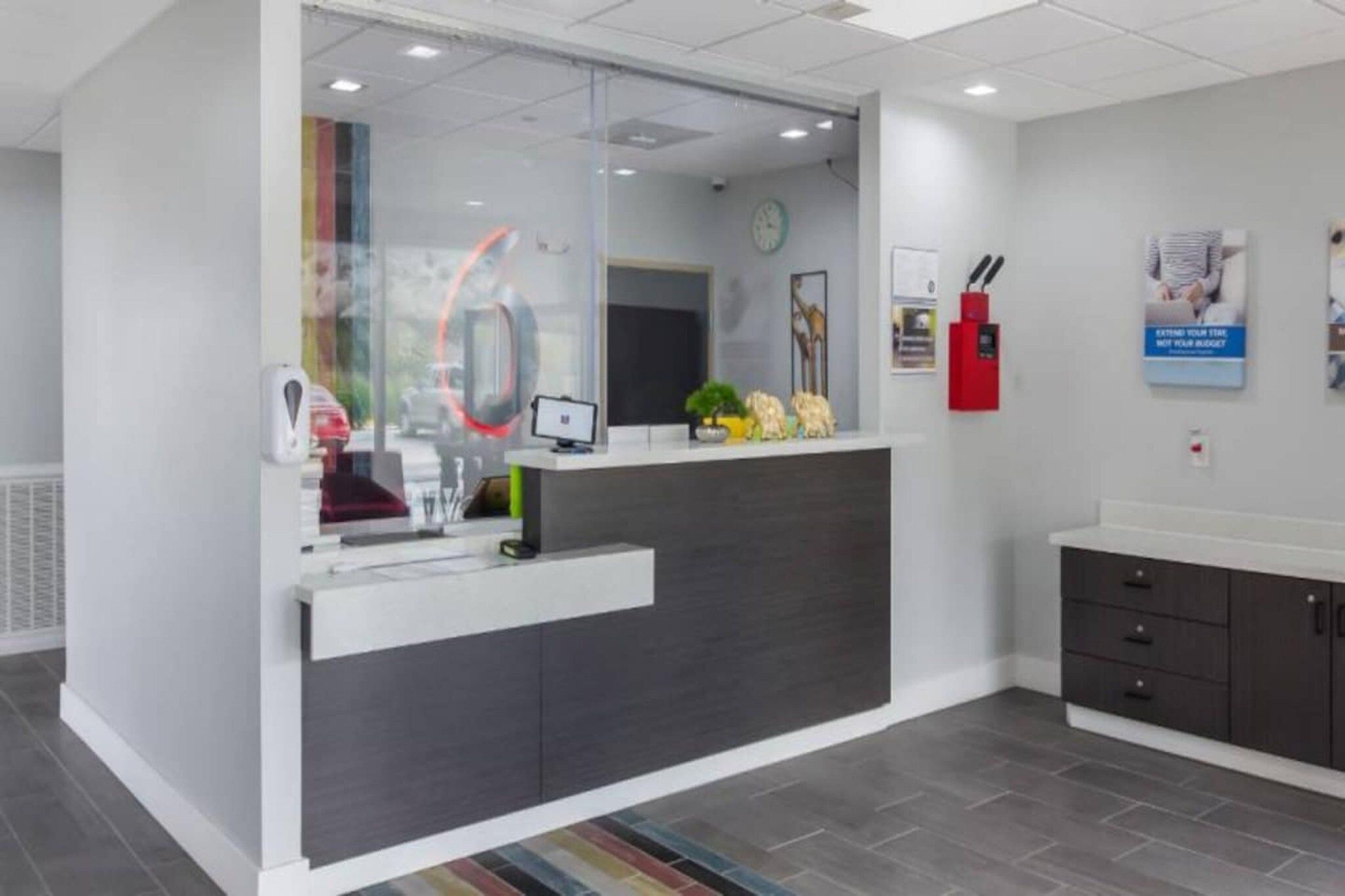 A reception area with a glass partition, a reception desk, a clock, a fire extinguisher, and a poster on the wall.