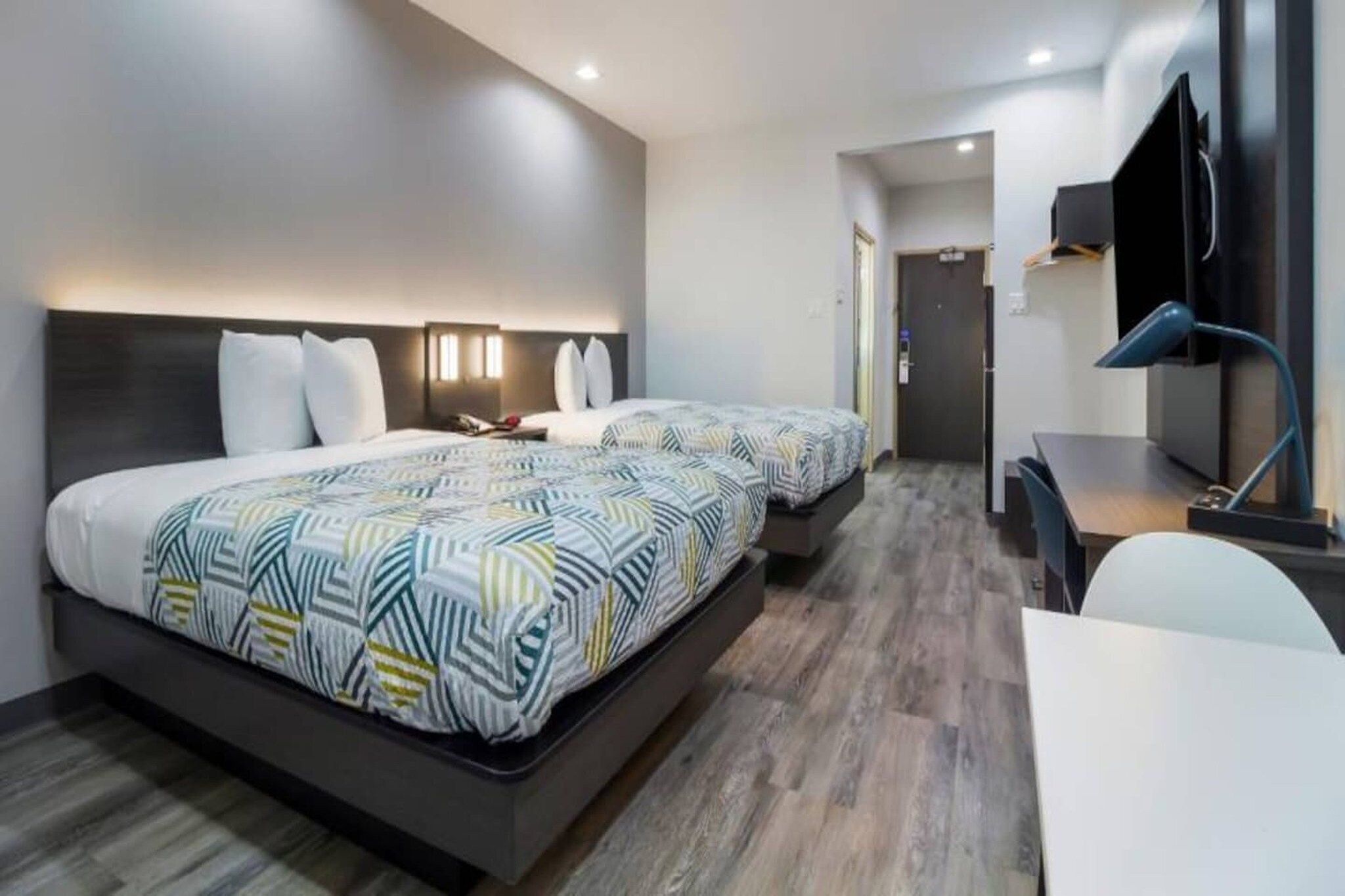 A hotel room with two beds, a desk, a chair, a TV, and a door leading to a hallway.