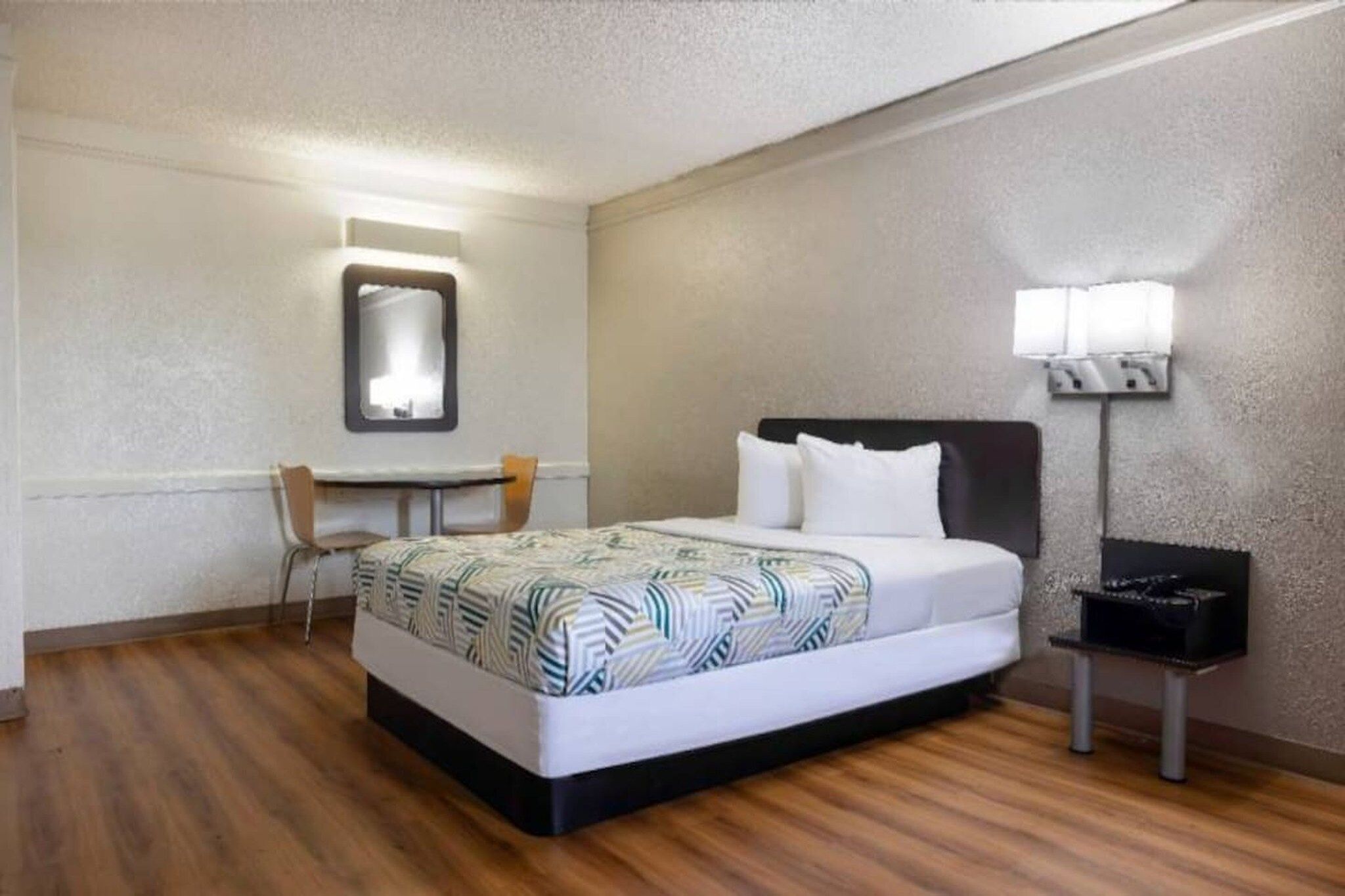 A hotel room with a bed, a desk, a chair, a nightstand, and a wall-mounted lamp.
