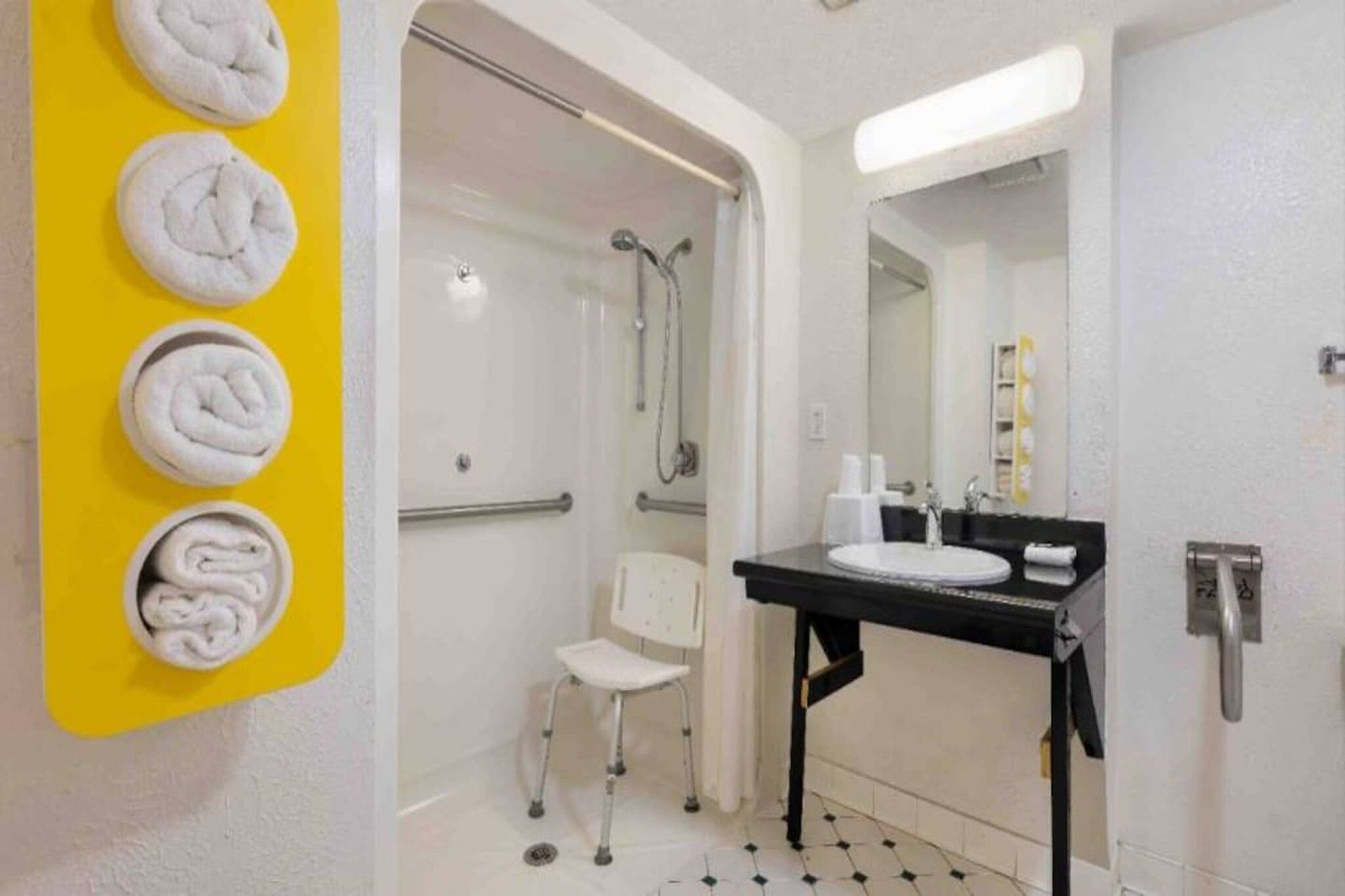 Un baño con ducha, lavamanos, una silla y toallas sobre una pared amarilla.