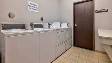 A laundry room with multiple washing machines, a sign stating 'Motel 6 is not responsible for items left unattended', and a 'MAINTENANCE' door.