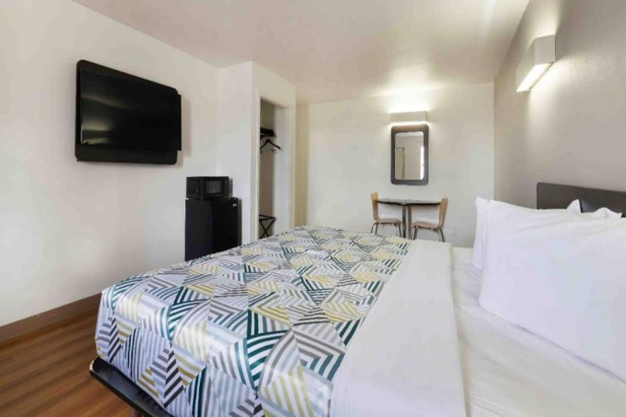 A hotel room with a bed, a flat-screen TV mounted on the wall, a desk with a chair, and a mirror.