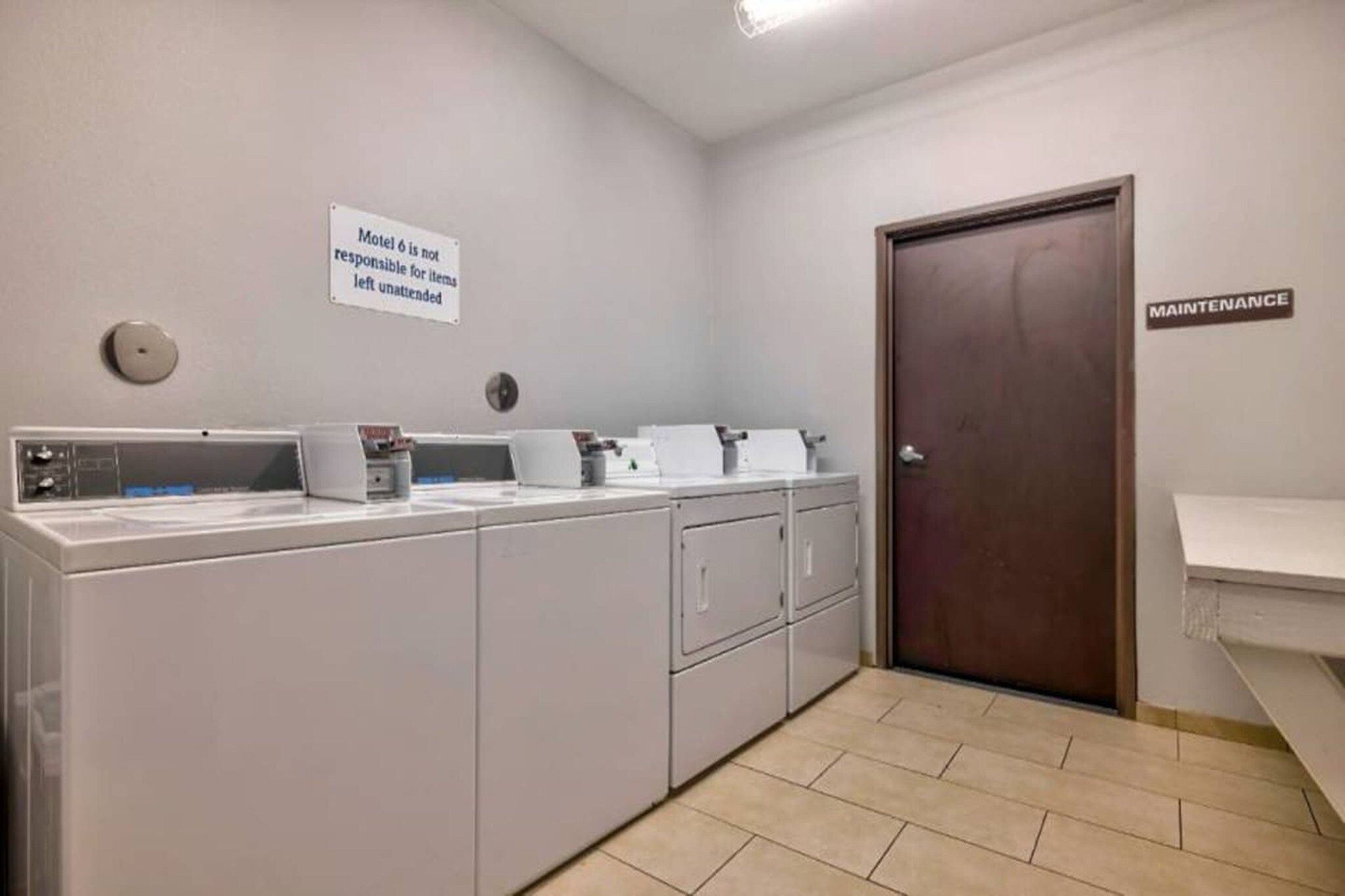 A laundry room with stacked washers and dryers, a sign stating 'Motel 6 is not responsible for items left unattended', and a 'MAINTENANCE' sign on the wall.