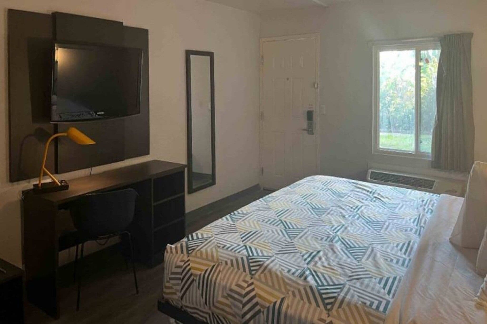 Hotellihuone, jossa on sänky, työpöytä, tuoli, televisio, peili, ovi ja ikkuna.