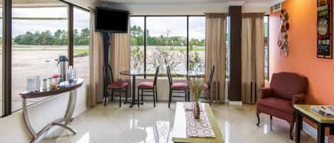 A hotel room with a large window, a flat-screen TV, a dining table with chairs, and a red armchair.