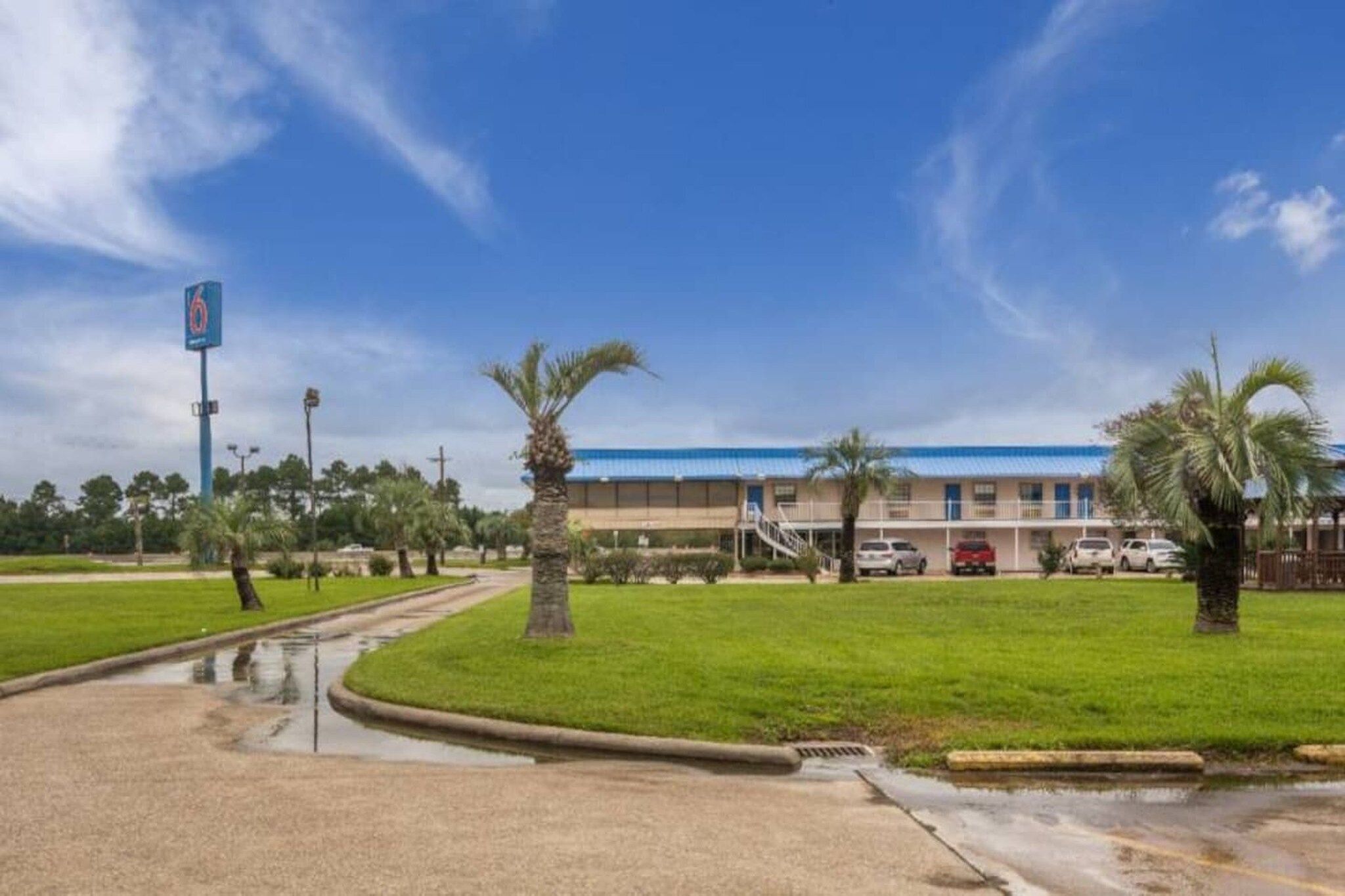 A hotel with a flat roof, a parking lot, and a grassy area with palm trees.