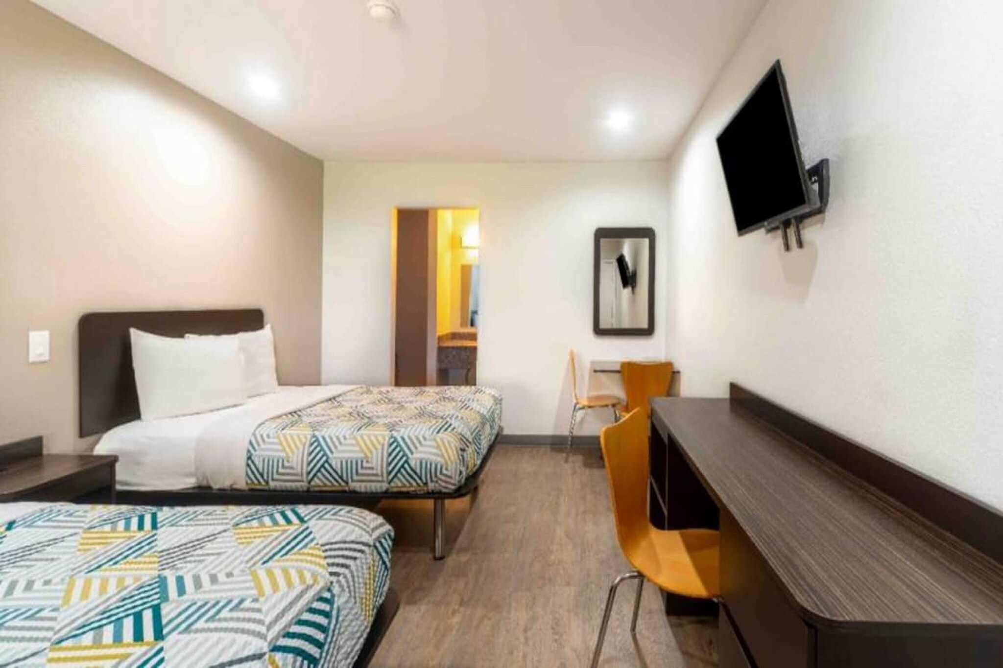 Habitación de hotel con dos camas, un escritorio, una silla, un televisor y una puerta que da a otra habitación.