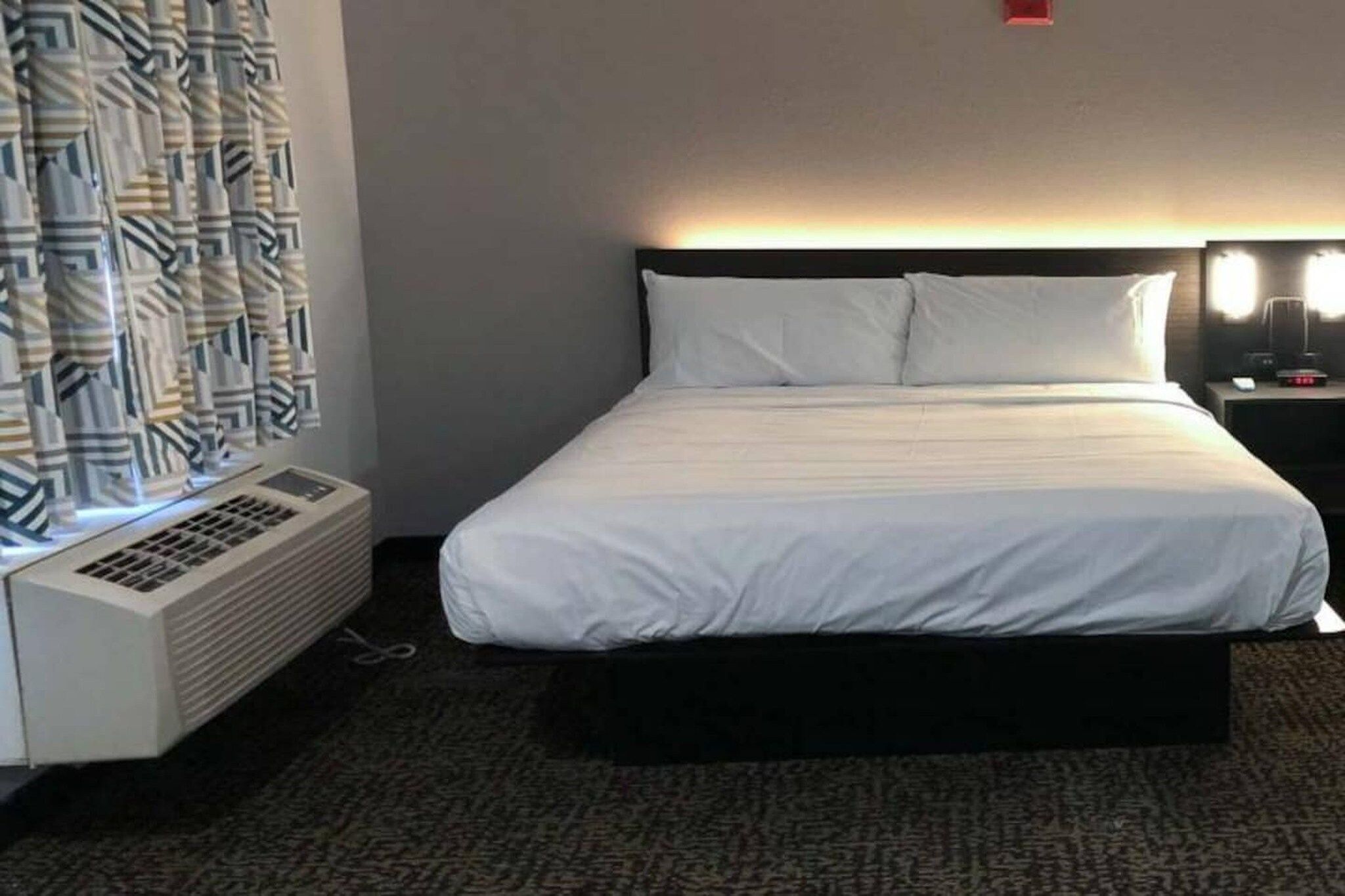 A hotel room with a bed, a heater, a nightstand with a lamp, and a patterned curtain.