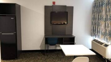 A compact room with a desk, a chair, a small table, a refrigerator, and an air conditioning unit.