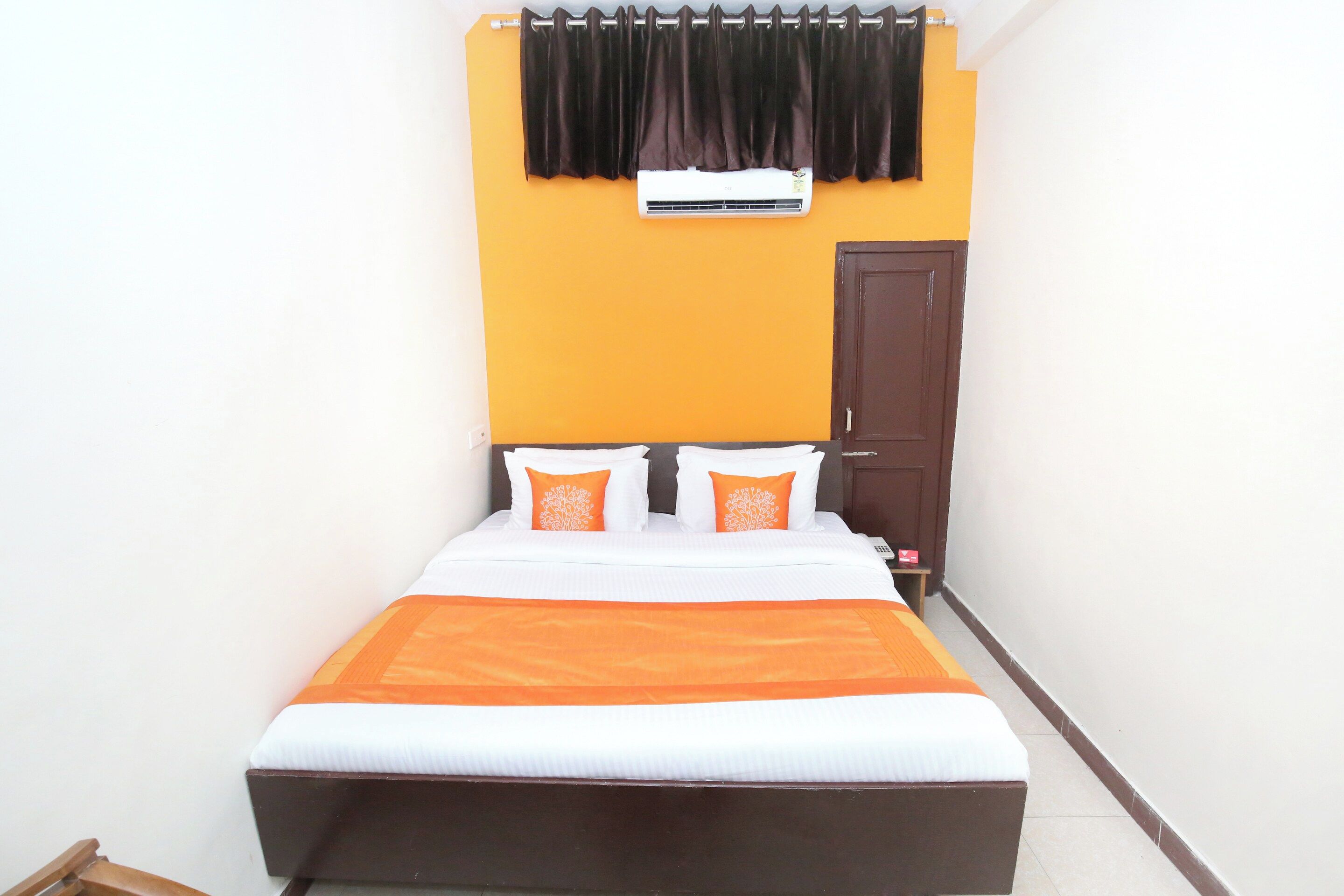 A small, narrow hotel room with a single bed, an air conditioning unit above the bed, and a dark-colored wardrobe.