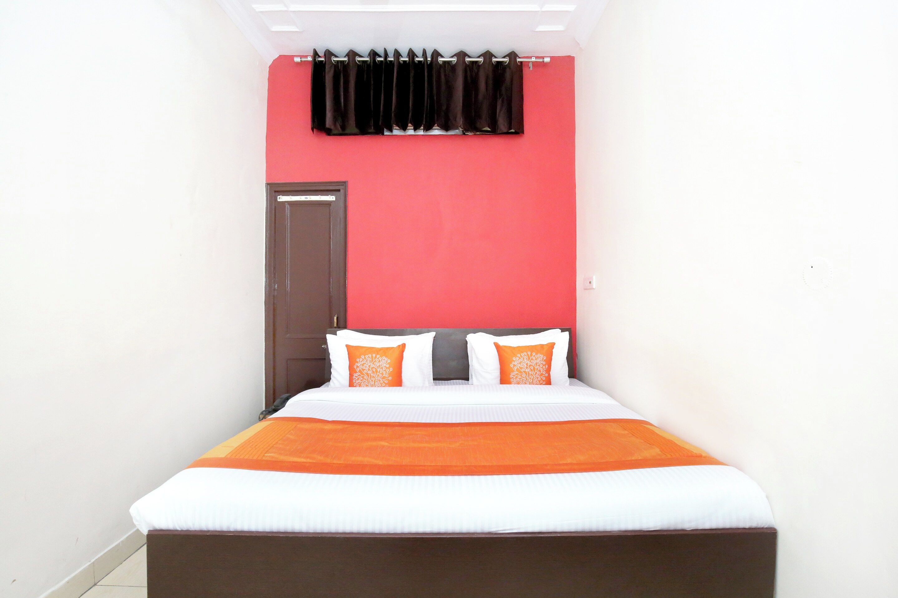 A narrow hotel room with a bed, two pillows, a red wall, and a black curtain.