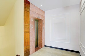 A hallway with a wooden panel wall, a marble wall with a lift sign, and a white door with a geometric design. - Collection O Railway Station Dehradun (Dehradun)