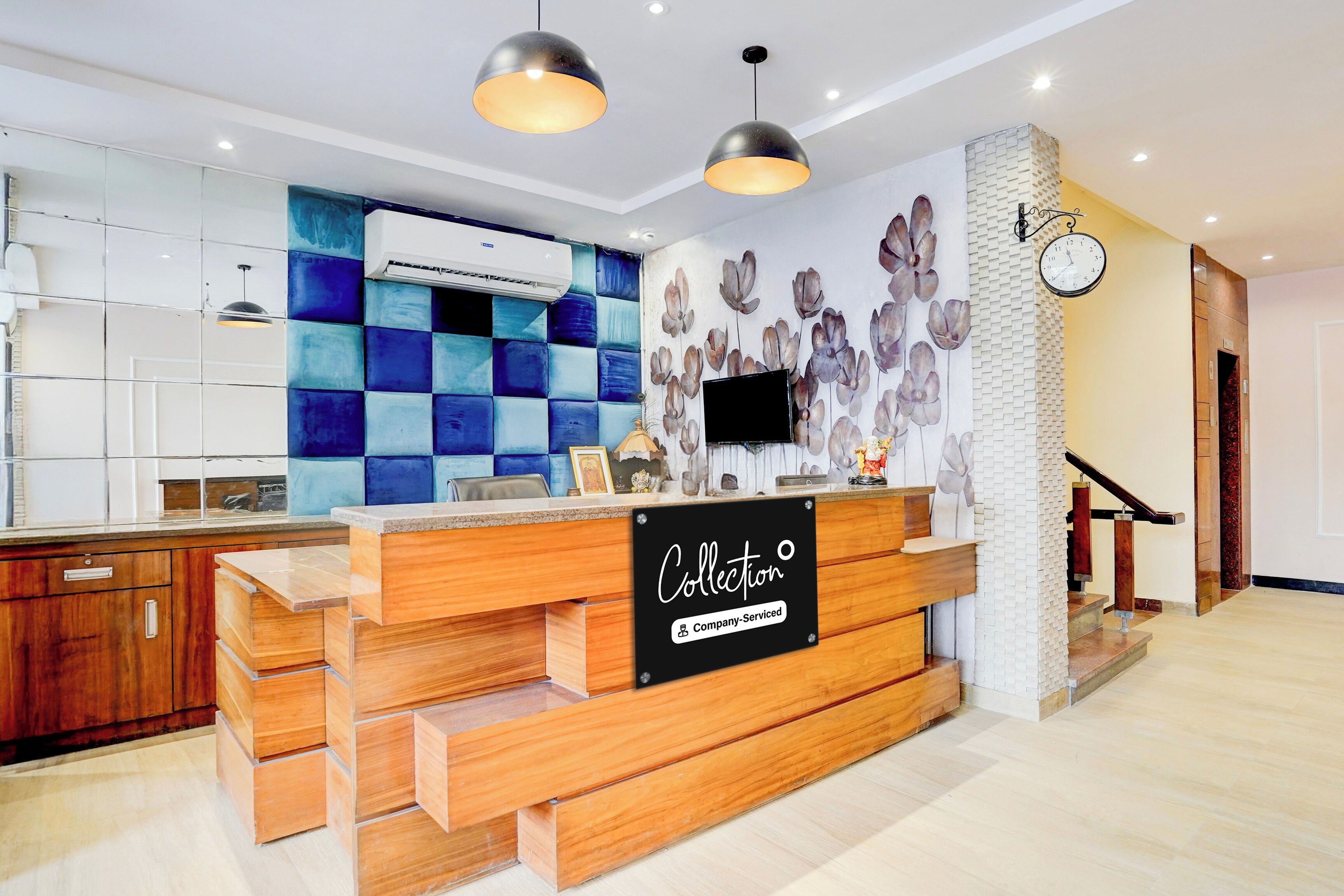 A reception desk with a sign indicating it's company-serviced, a wall-mounted clock, and a modern kitchen area with blue tiles and a large air conditioning unit.