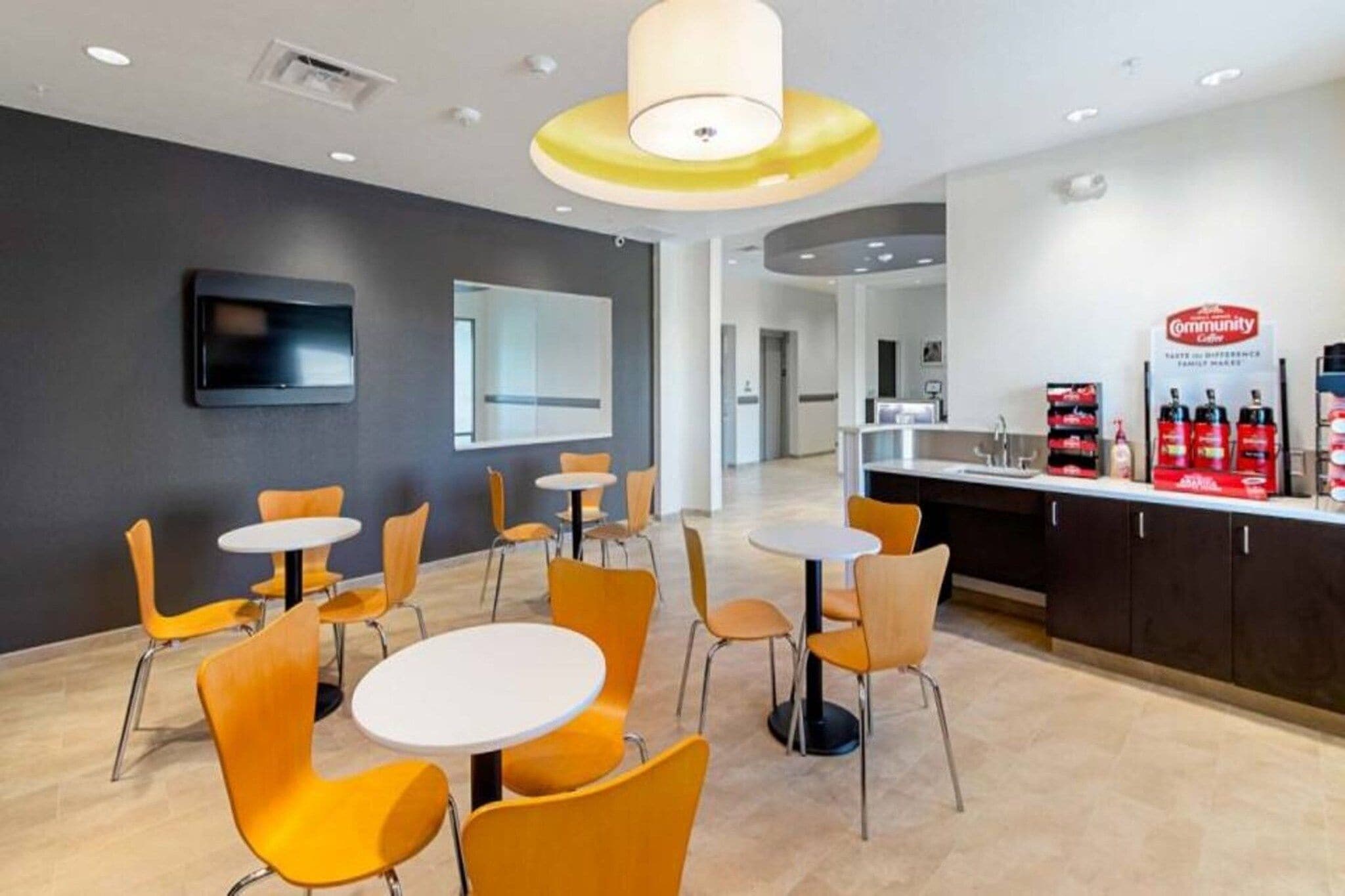 A modern interior with round tables and orange chairs, a flat-screen TV, and a coffee station.
