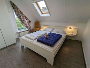Ein Schlafzimmer mit Dachfenster, einem Bett mit blauer Decke und einem Teddybären, einem Stuhl, einem Nachttisch mit Lampe und einem Fenster mit Vorhängen.