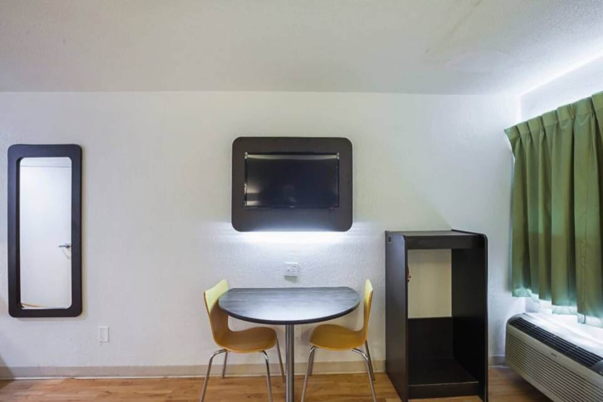 A small room with a round table and two chairs, a wall-mounted TV, a heater, and a window with curtains.