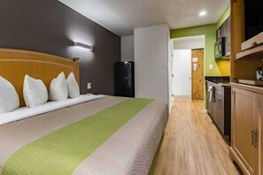 A hotel room with a bed, a refrigerator, a kitchenette, and a door leading to another room. - 1 King Bed | Non-Smoking, Kitchen (Plano)