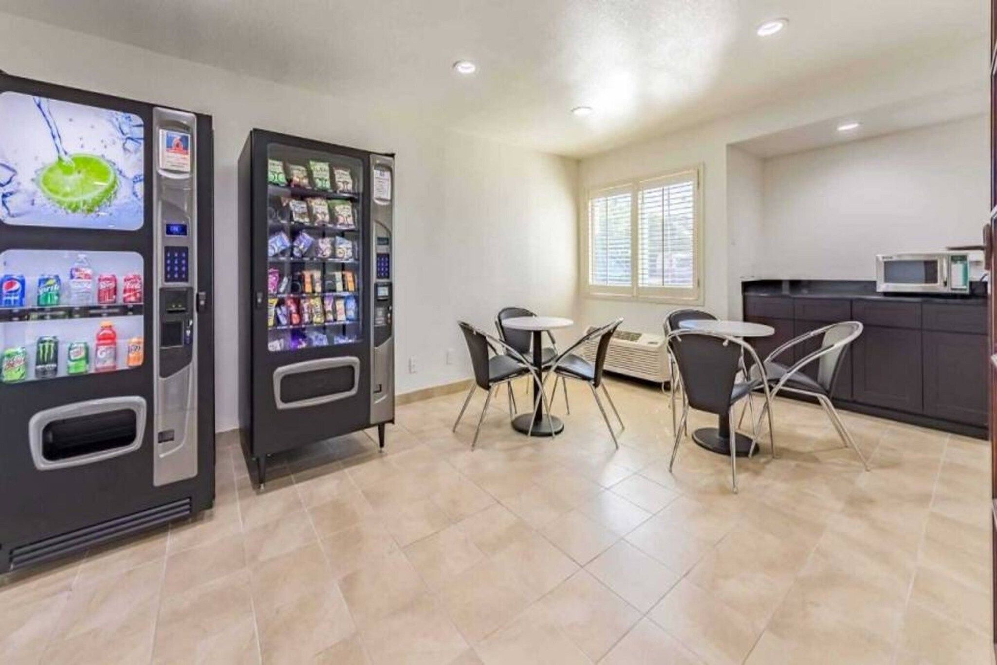 A clean, modern kitchen with two beverage vending machines, a dining area with four chairs, and a microwave on the counter.