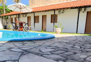 Une piscine avec un patio en pierre, une table entourée de chaises et un bâtiment doté d’un toit en tuiles.