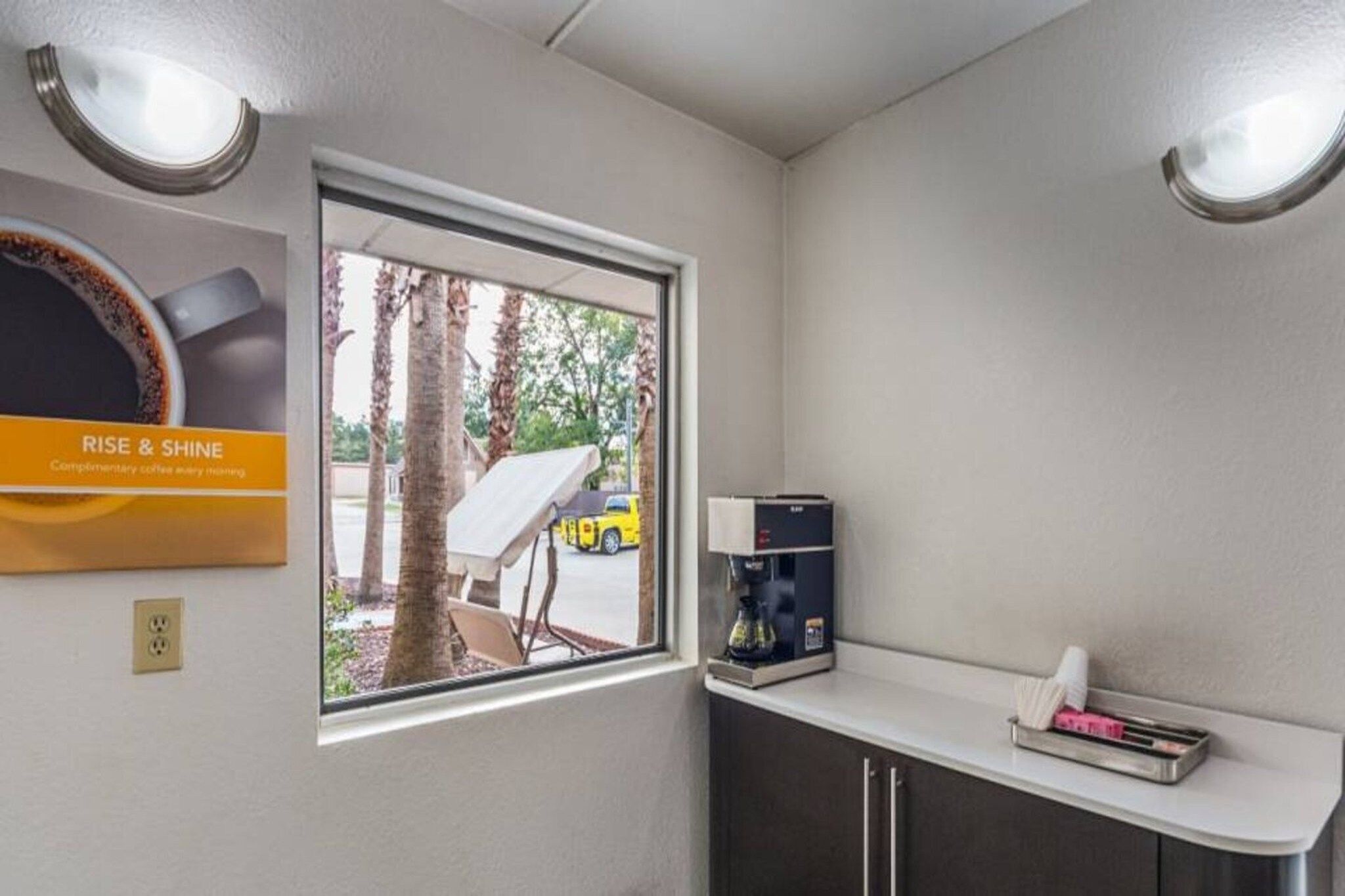A small kitchenette with a window, a coffee machine, and a sign that reads 'RISE & SHINE'.