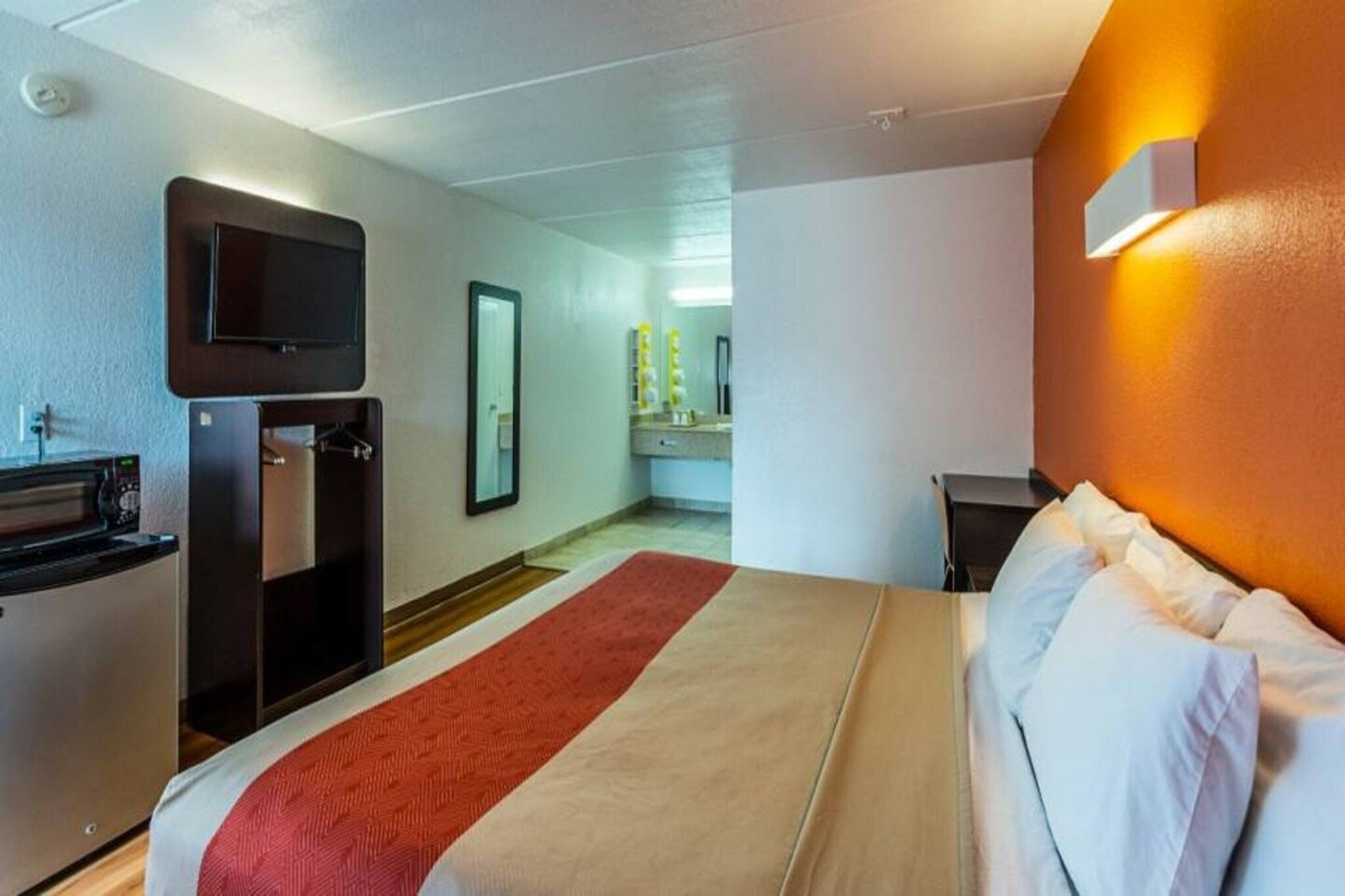 A hotel room with a bed, a desk with a computer, a mirror, and a bathroom visible through an open door.