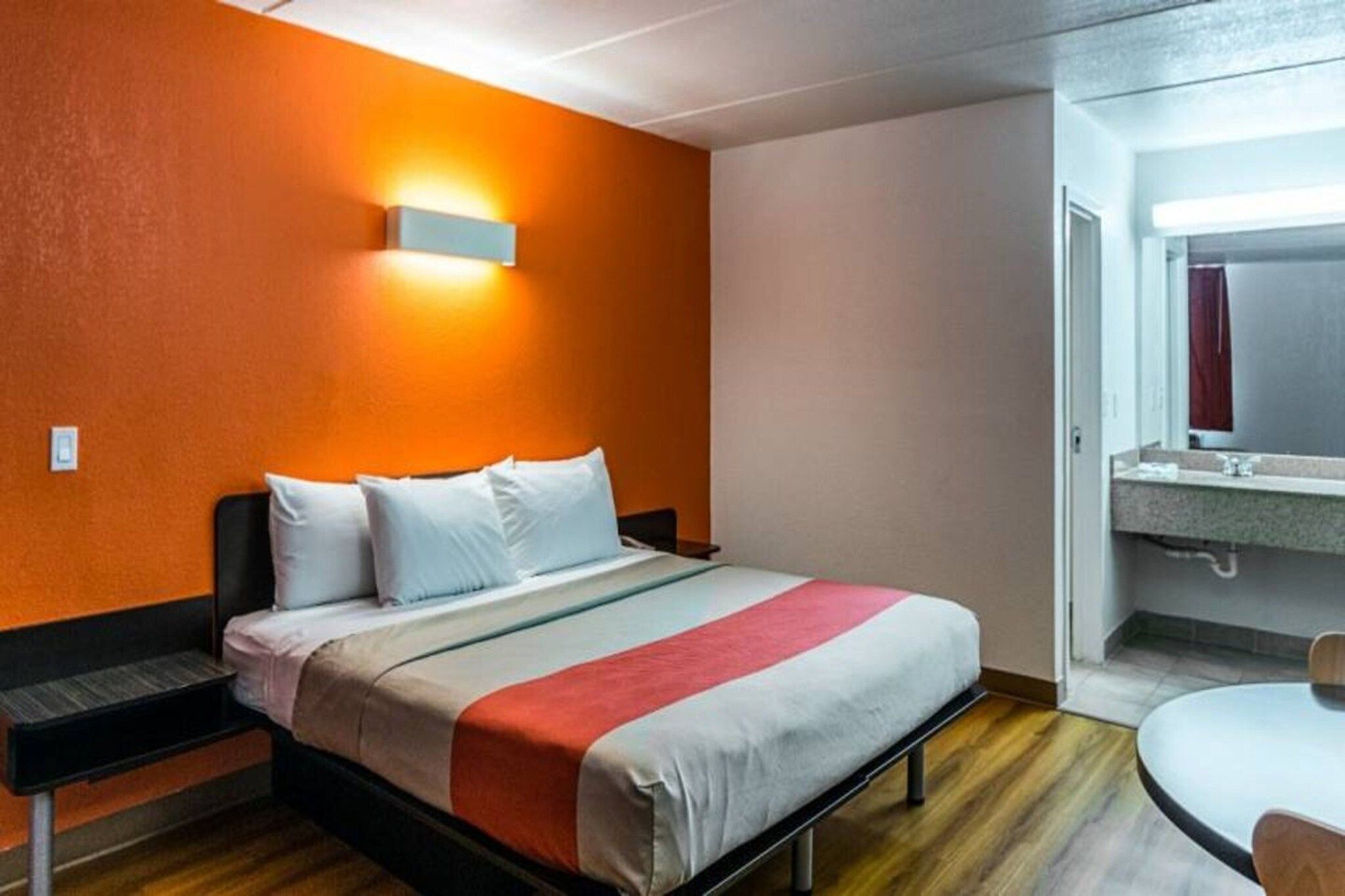 Une chambre d’hôtel comprenant un lit, une table de chevet, une lampe fixée au mur et une salle de bain avec un lavabo et un miroir.