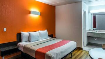 A hotel room with a bed, a nightstand, a wall-mounted light, and a bathroom with a sink and mirror.