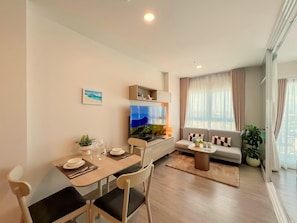 A modern living room with a dining area, a flat-screen TV, a sofa, and a large window with curtains. - New 1 BR Condo with Seaview (BW287) (Hua Hin)
